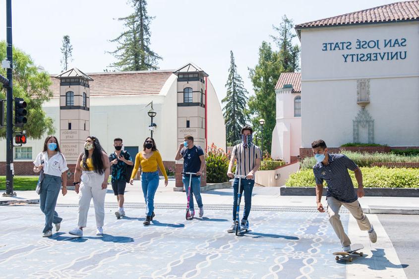 Students crossing the street and one skateboarding with masks on.